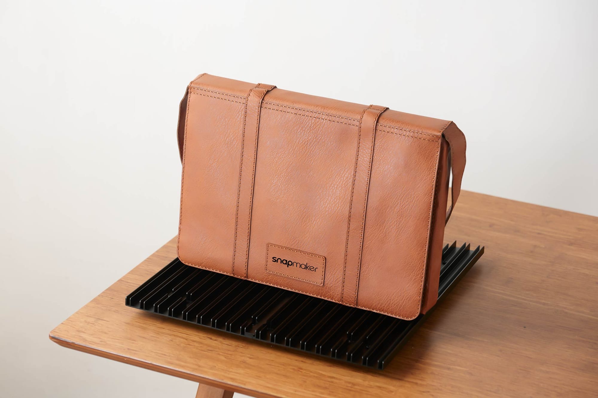 A brown leather messenger bag, displayed on a wooden table, features a custom laser-engraved patch with the "Snapmaker" brand logo on the front.