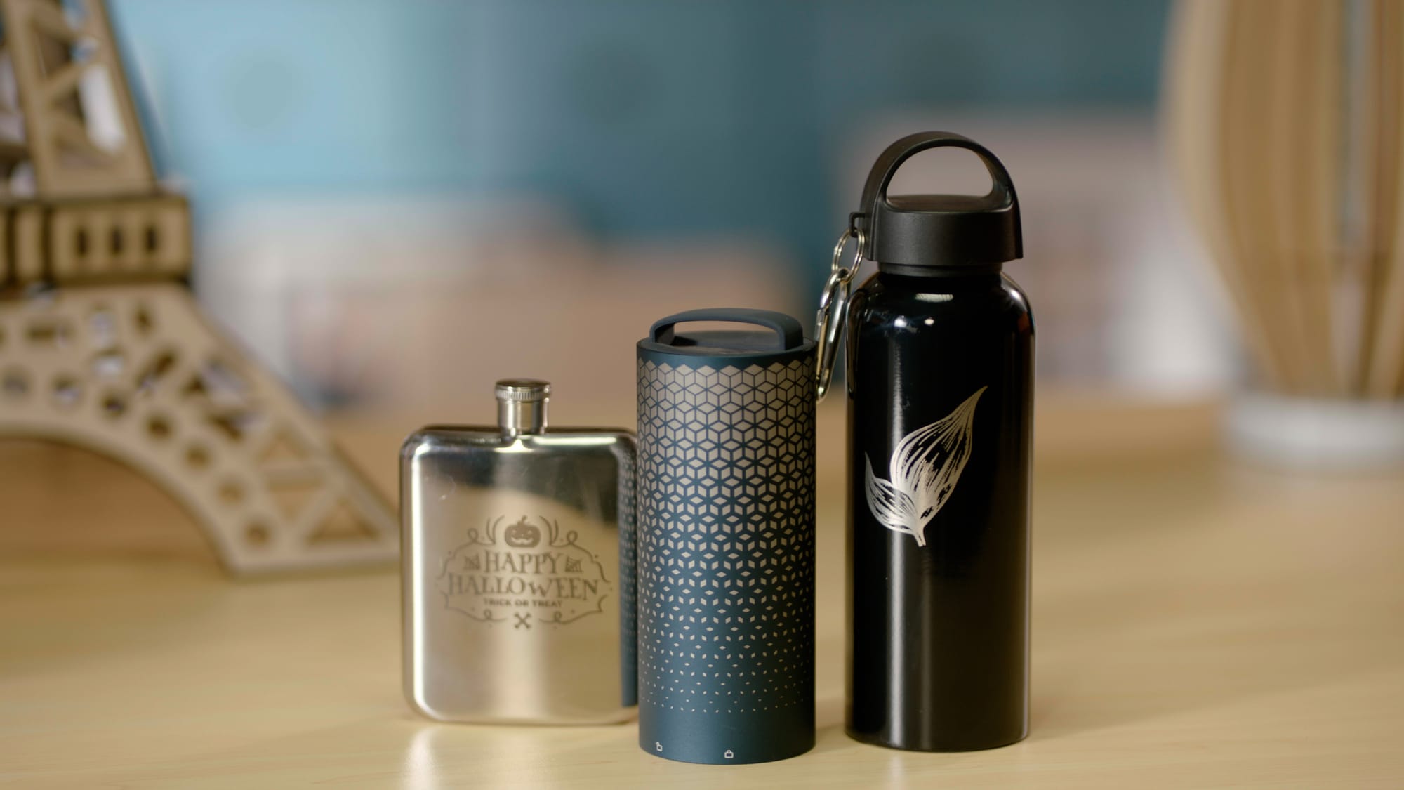 A silver flask, a patterned tumbler, and a black water bottle sit on a wooden table, each featuring a unique design created by a laser engraver.