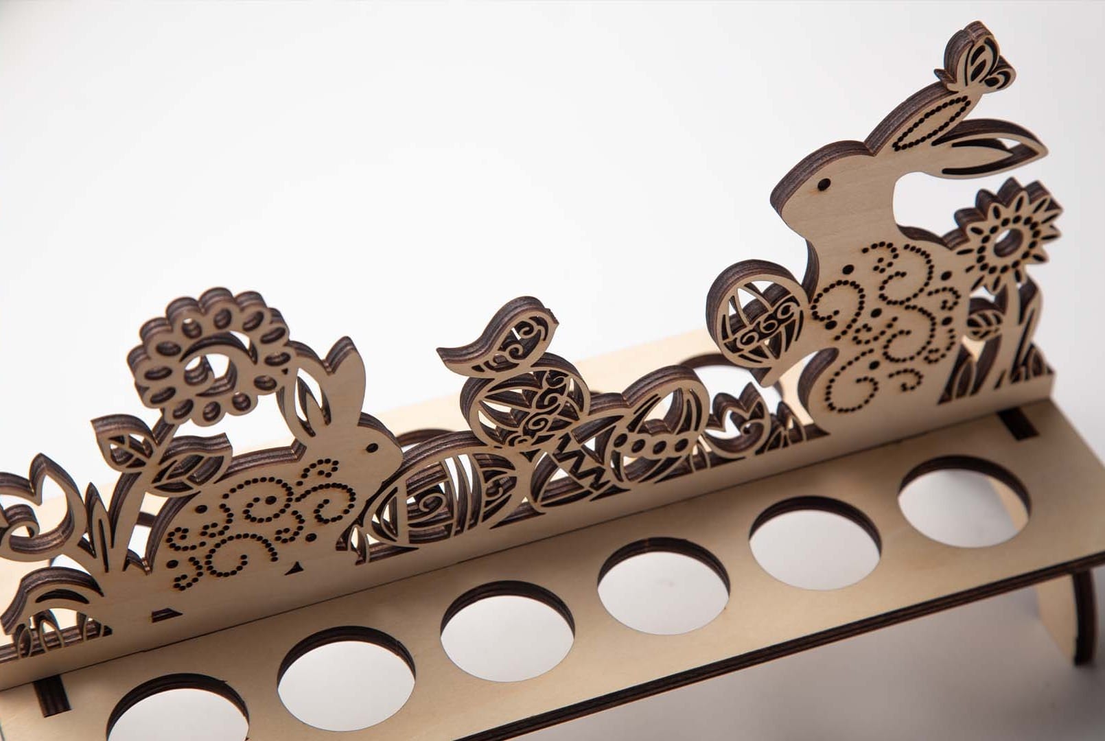 A close-up of an intricate, laser-cut wooden Easter egg holder featuring decorative cutouts of bunnies, chicks, and flowers, against a clean white background.