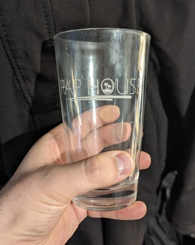 A person's hand holds up a clear drinking glass that has been laser-engraved with a logo featuring a palm tree.