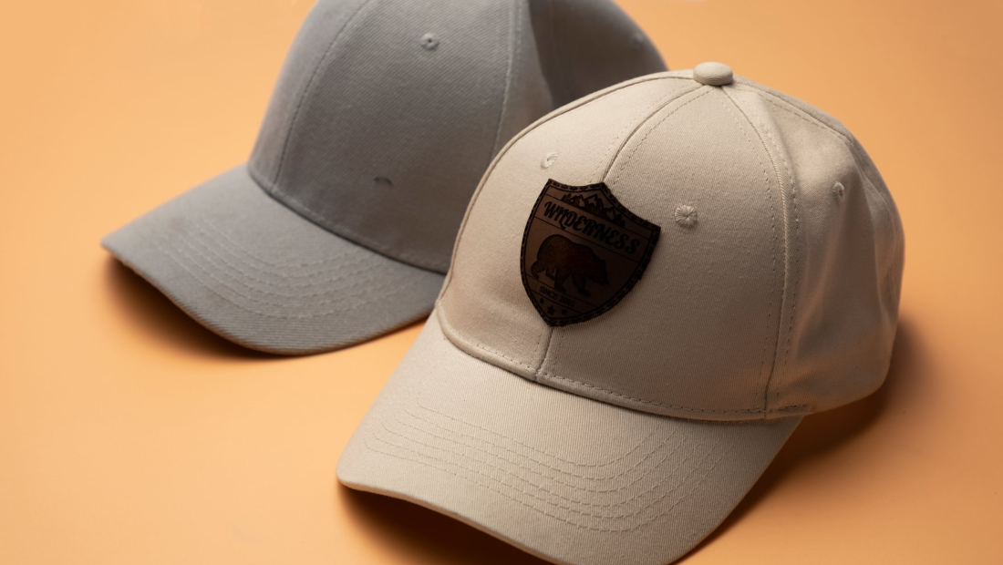 A beige baseball cap with a custom laser-engraved leather patch showing a bear and the word "Wilderness" sits in front of a plain gray cap.