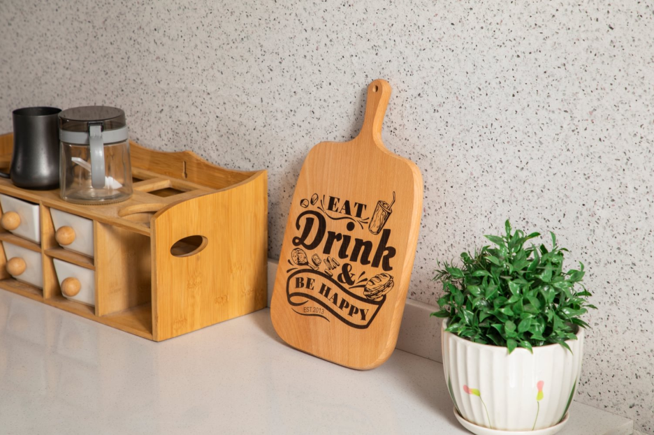 A laser-engraved wooden cutting board with the phrase "Eat Drink & Be Happy" sits on a kitchen counter next to a spice rack and a small potted plant.