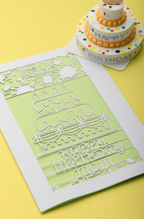 An intricate, laser-cut "Happy Birthday Mom" card featuring a cutout of a birthday cake sits on a yellow surface next to a small decorative cake.