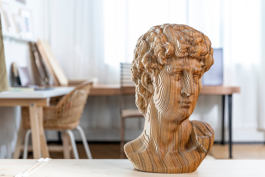 A 3D model of a classical bust, created from stacked layers of laser-cut wood, sits on a workbench in a well-lit studio.