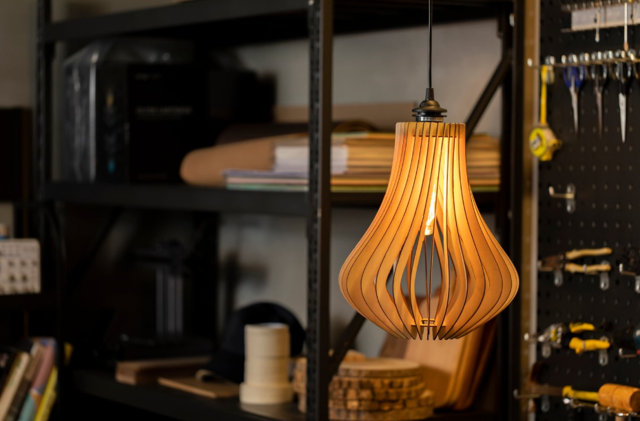 A glowing, laser-cut wooden pendant lamp with a curved slat design hangs in a workshop, with shelves and a tool-filled pegboard visible in the background.