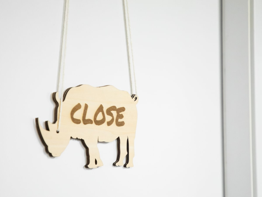 A laser-cut wooden business sign in the shape of a rhinoceros with the word "CLOSE" engraved on it hangs from a rope on a white door.
