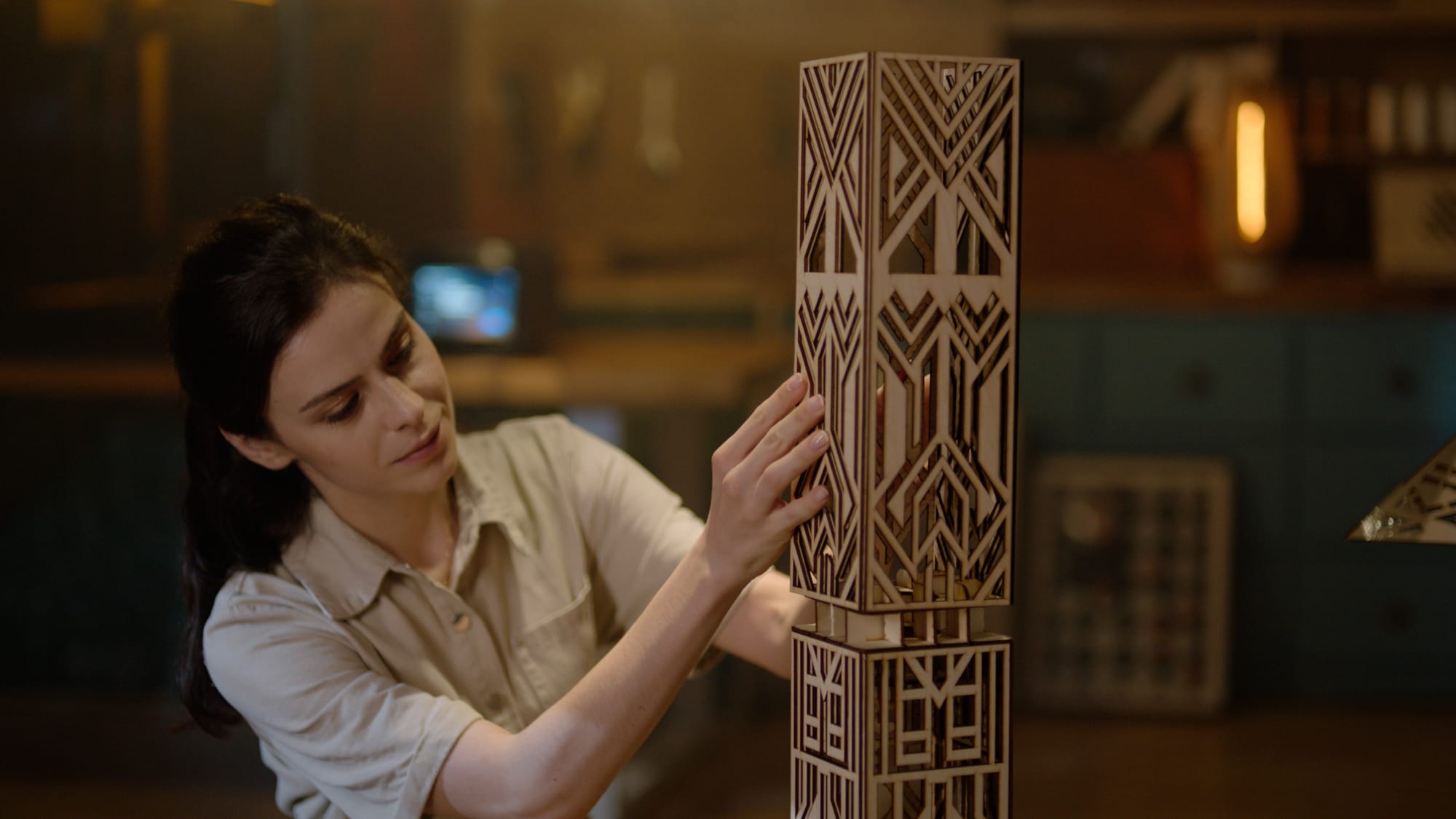 A woman in a workshop carefully assembles a tall, ornament made from intricately laser-cut wooden panels with a geometric, Art Deco-style pattern.
