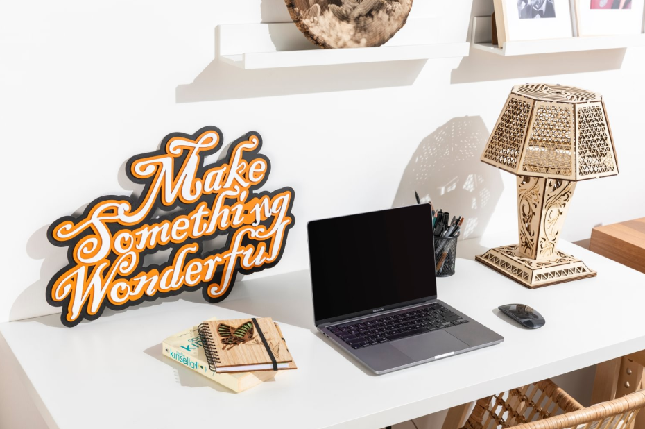 A white desk displaying custom laser-engraved items, including a 'Make Something Wonderful' sign, an intricate wooden lamp, and a butterfly-design notebook next to a laptop.