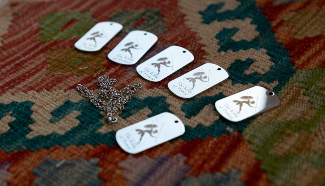 Several laser-engraved, silver dog tags featuring a warrior graphic and the text "The King of Power" are displayed on a colorful patterned rug with a ball chain.