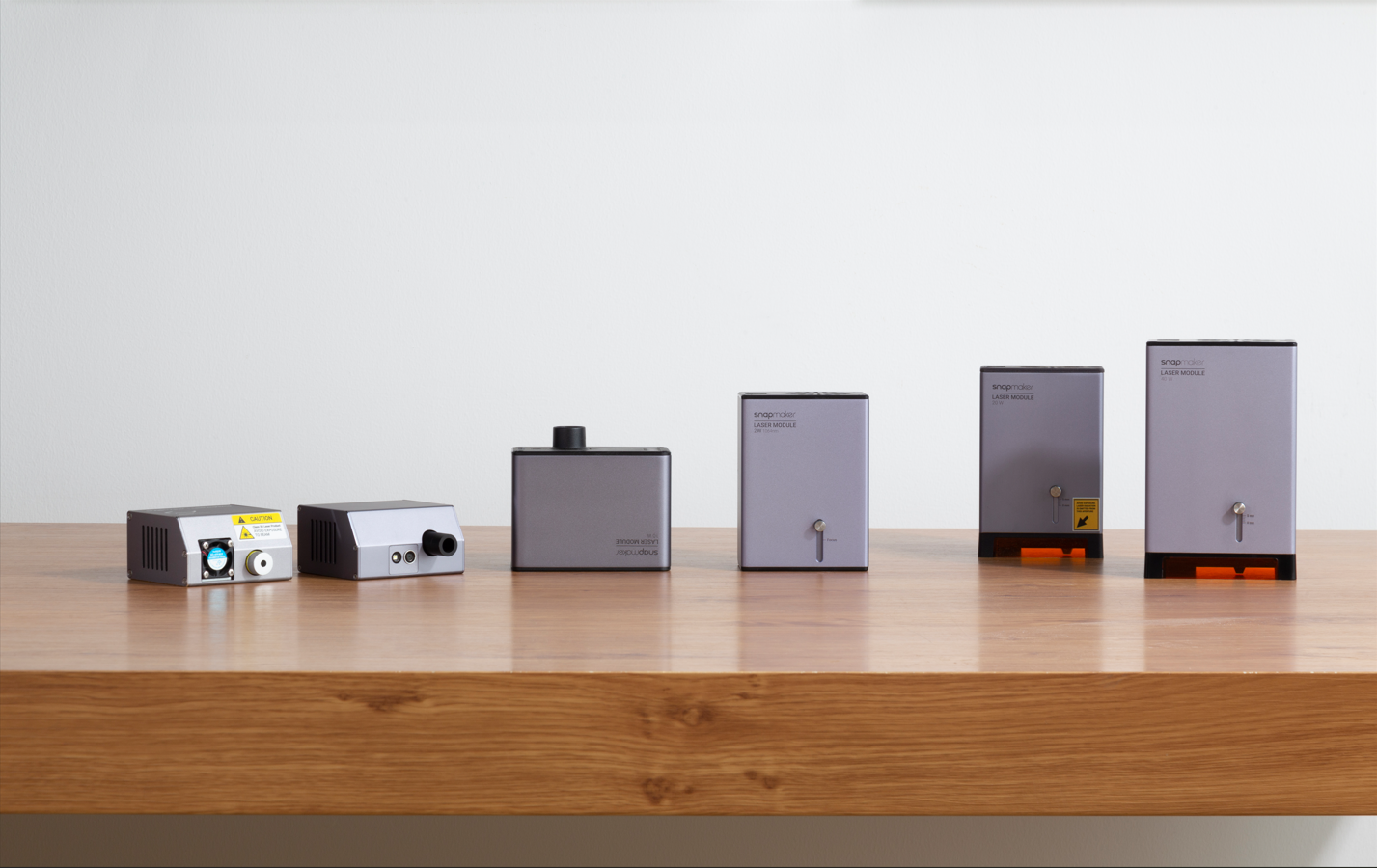 A side-by-side lineup of five different Snapmaker laser modules on a wooden surface, showing the progression in design and power.