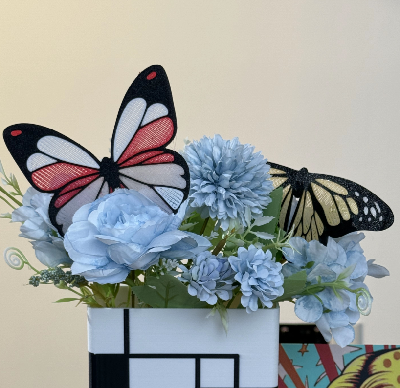 A close-up of two multi-material 3D printed butterflies with delicate, colorful wings arranged decoratively in a vase with blue flowers.