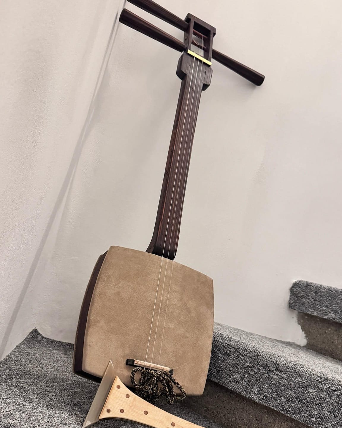 A three-stringed Japanese shamisen with a carved wooden body leans against a white wall and staircase.
