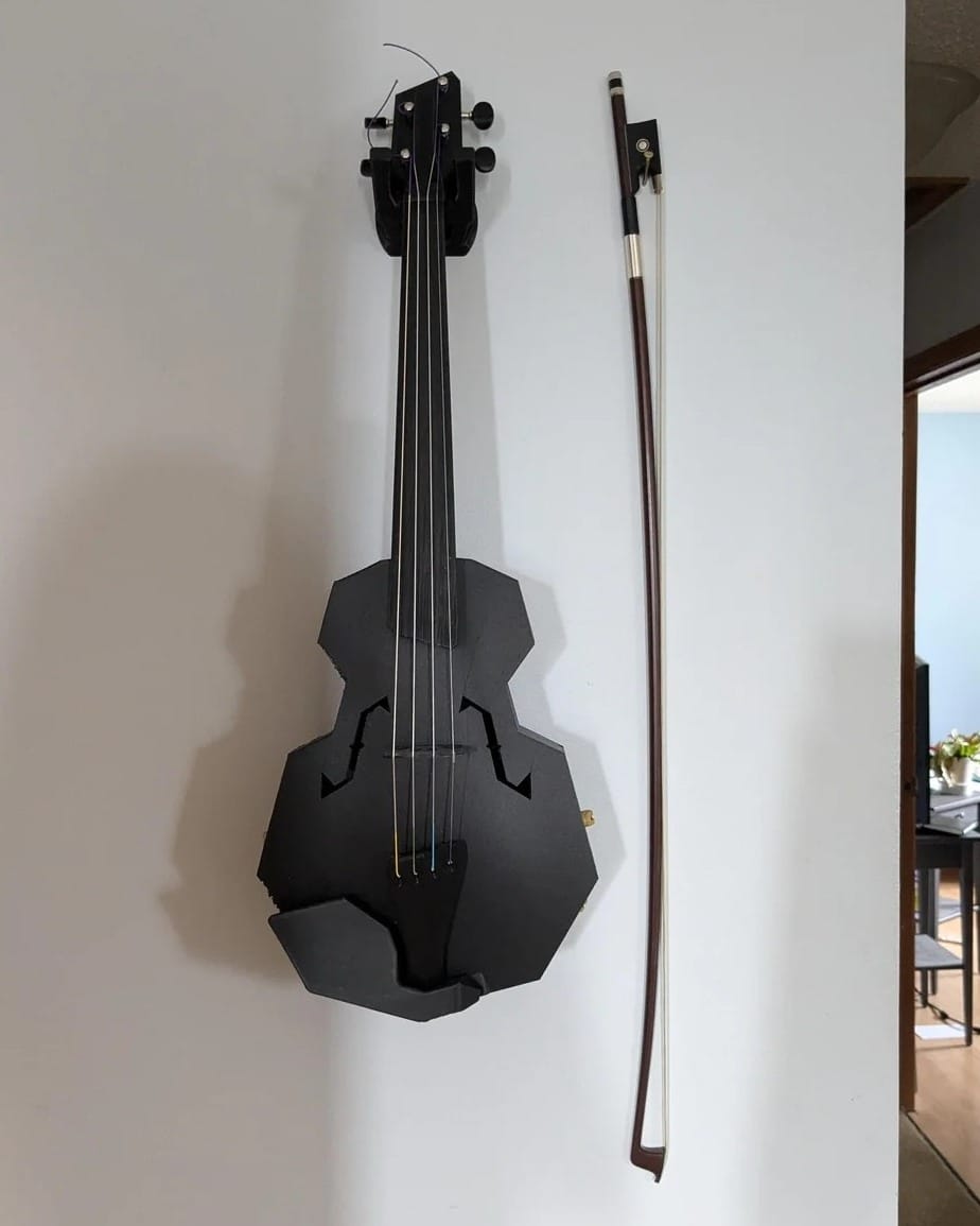 A full-size 3D-printed black violin with a bow hangs on a white wall.