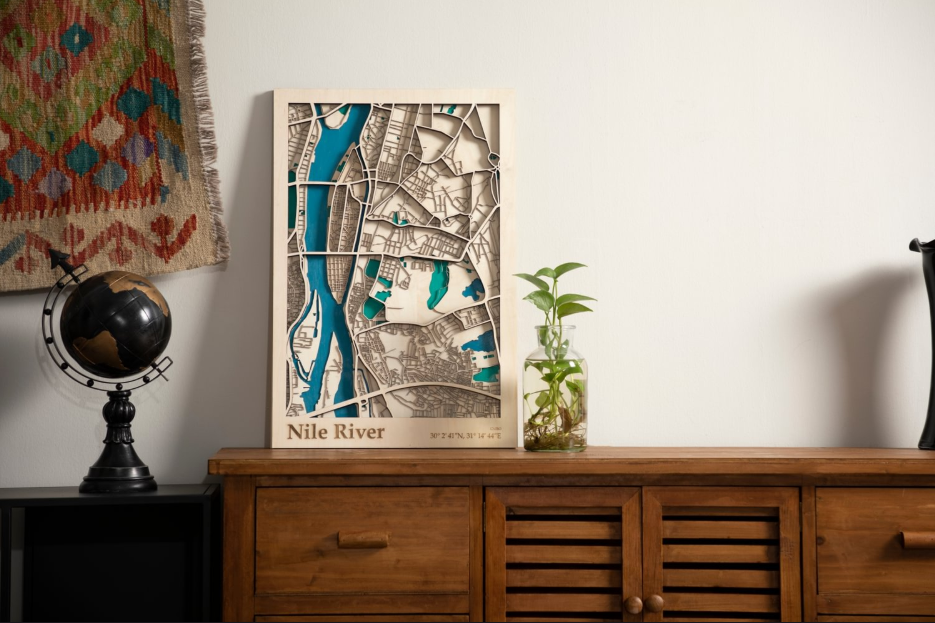 A close-up shot of a laser-engraved, multi-layered wooden map of the Nile River, displayed on a wooden cabinet with a globe and a plant in a glass vase nearby.