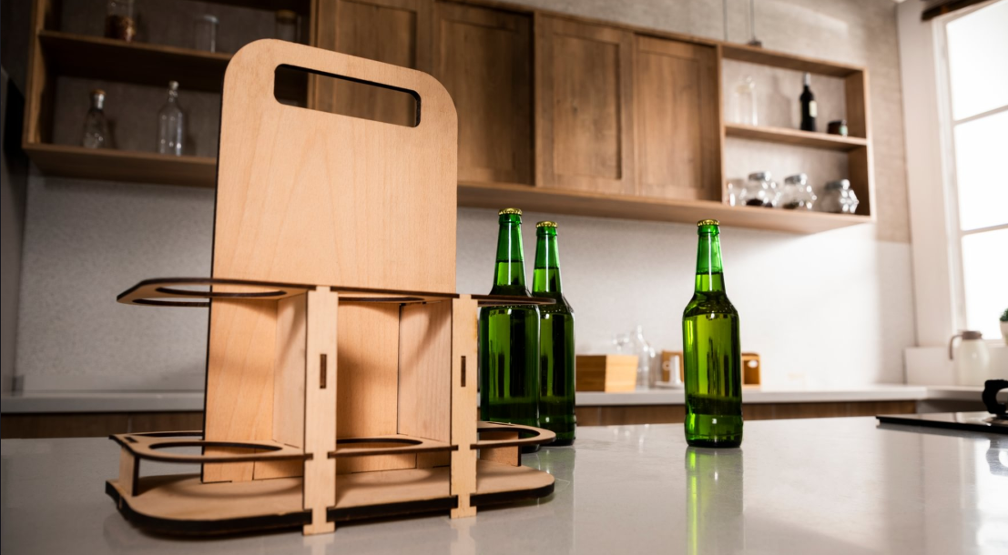A laser-cut wooden six-pack beer caddy with a carrying handle sits on a modern kitchen counter next to three green glass bottles.