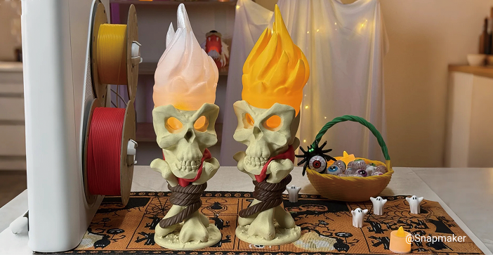 Two functional 3D printed skull torches with glowing flames, displayed on a Halloween-themed table runner next to 3D printer filament spools and a basket of treats.