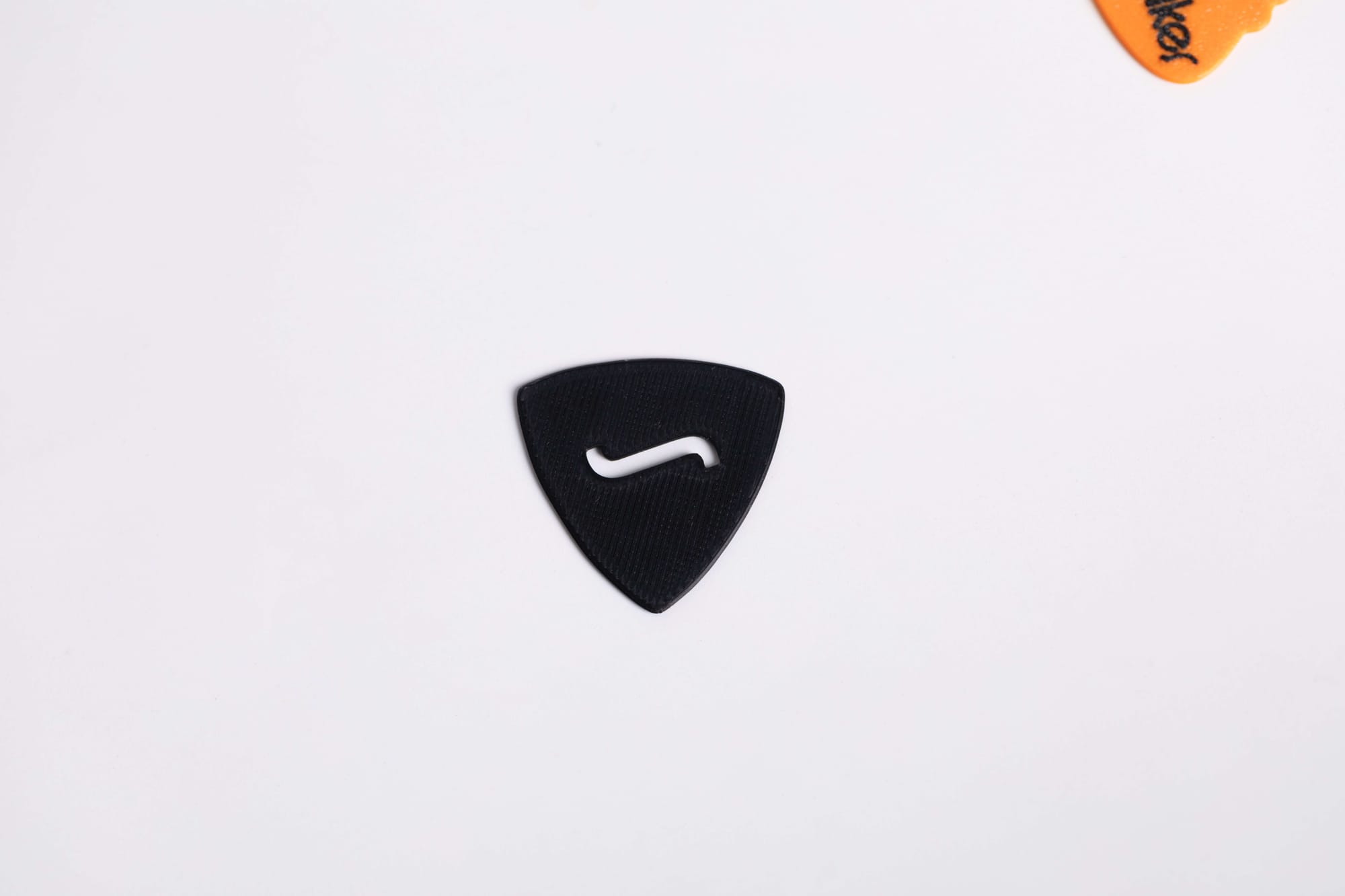 A single black, triangular 3D-printed guitar pick with a cut-out 'S' shape in the middle, resting on a clean white surface.