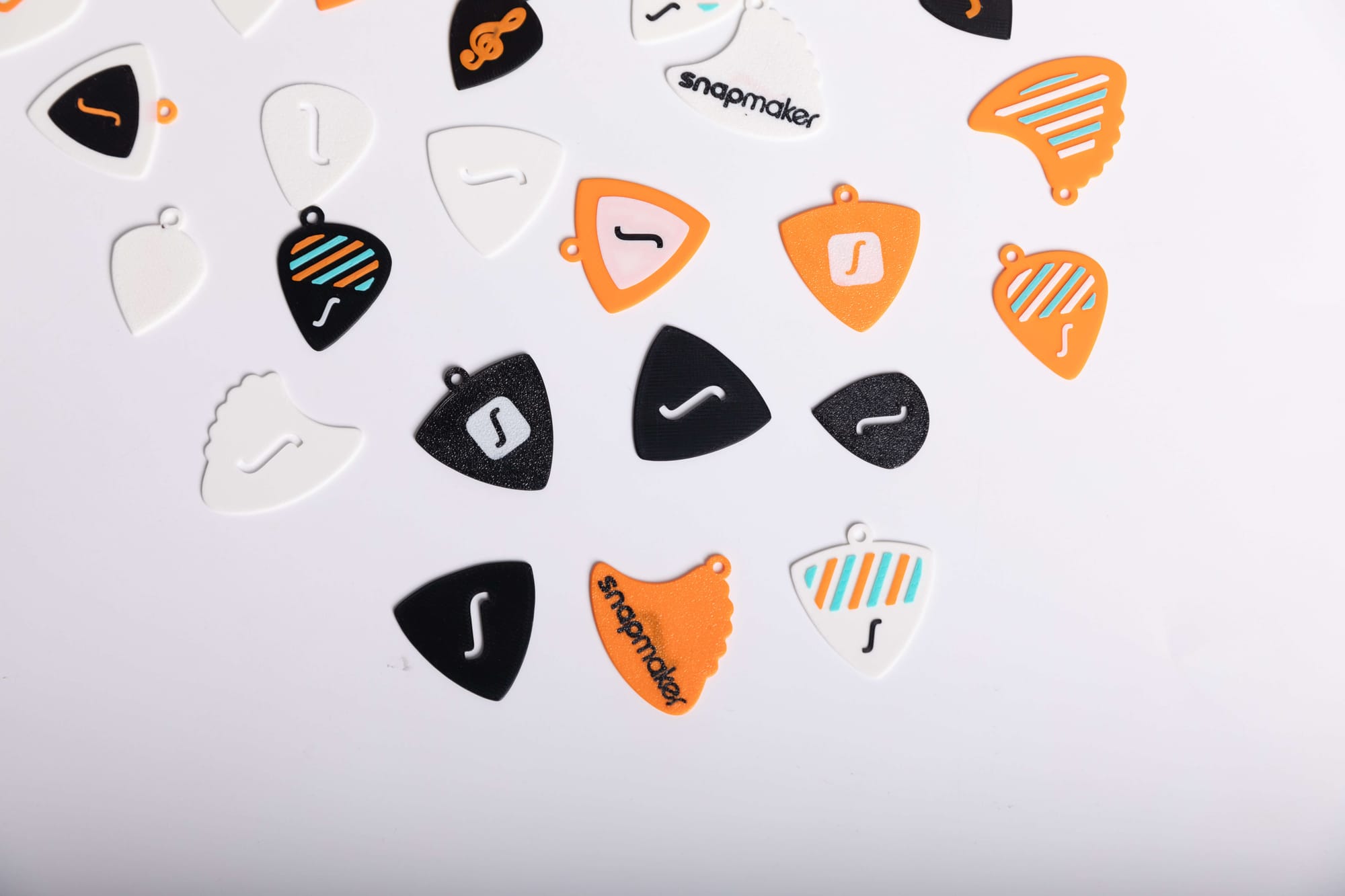 A close-up view of several 3D-printed guitar picks in various colors and designs, including black, white, and orange, with some featuring stripes or the Snapmaker logo.