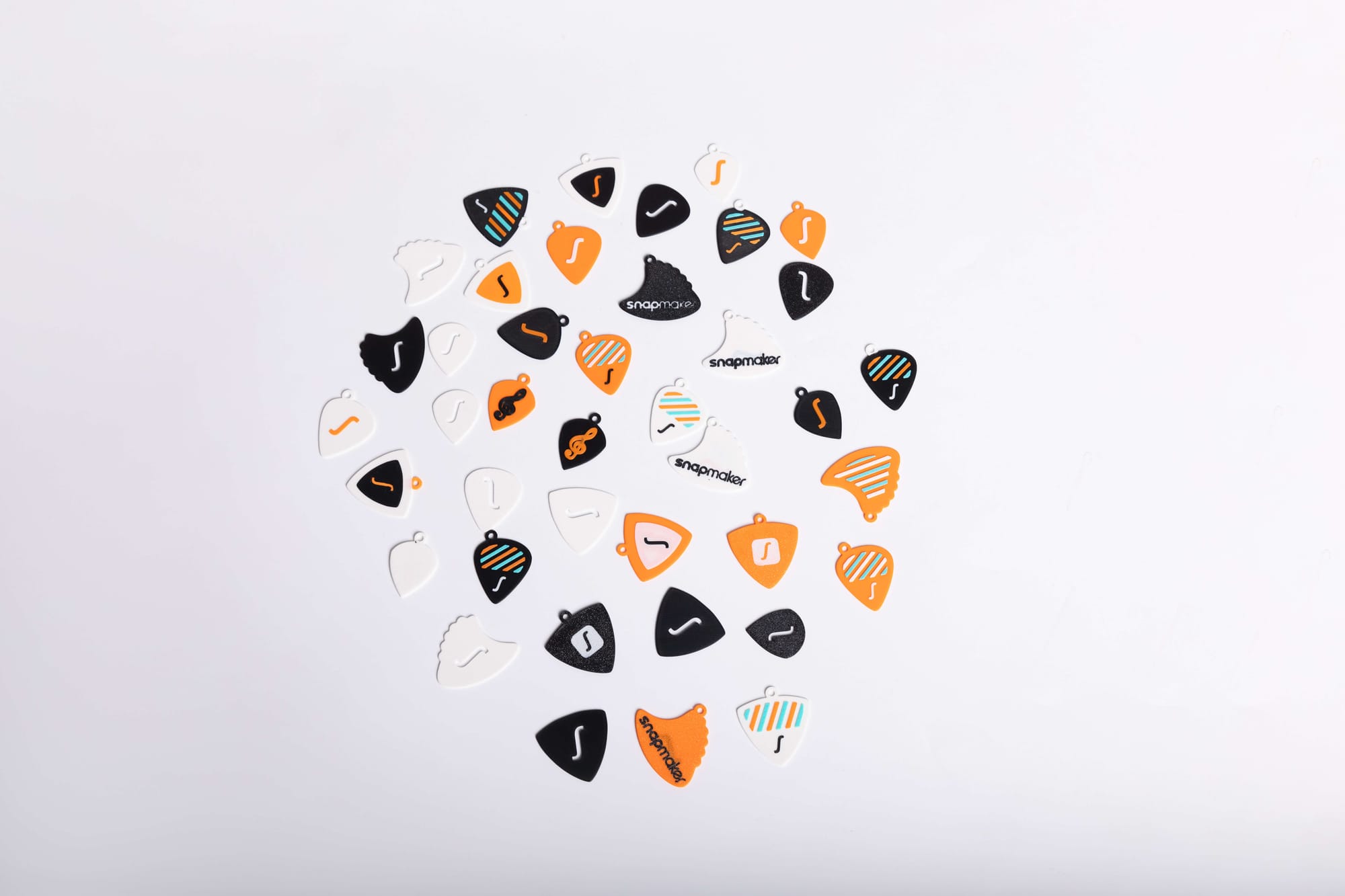 An overhead shot showing a variety of custom 3D-printed guitar picks, some with unique textures, patterns, and the Snapmaker brand name, spread across a white surface.