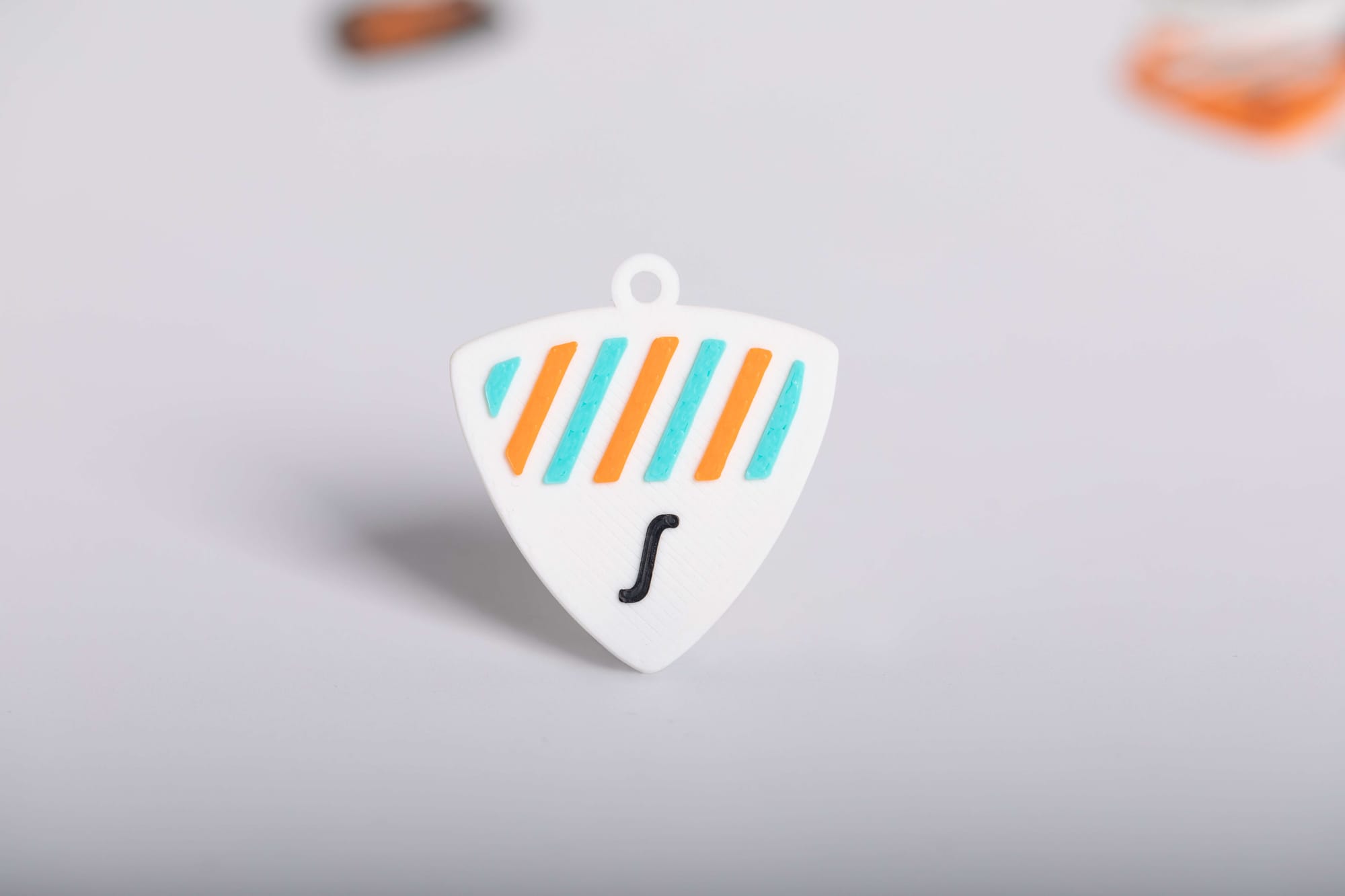 A single white, shield-shaped 3D-printed guitar pick with orange and teal stripes and a black 'S' logo in the center, featuring a small loop for a keychain.