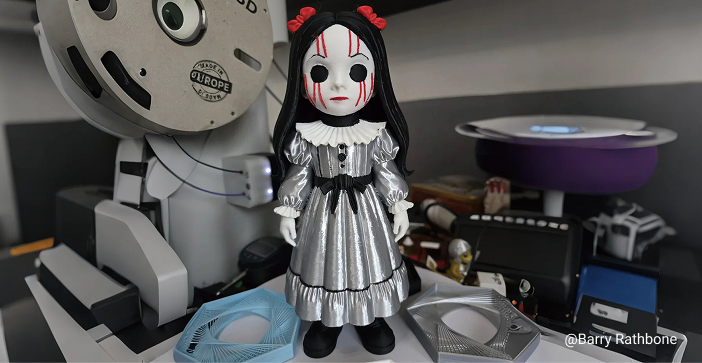 A highly detailed 3D printed doll figurine with a porcelain-like finish, white face with black eyes and red markings, wearing a shiny silver dress and black boots, standing on a desk with machinery in the background.