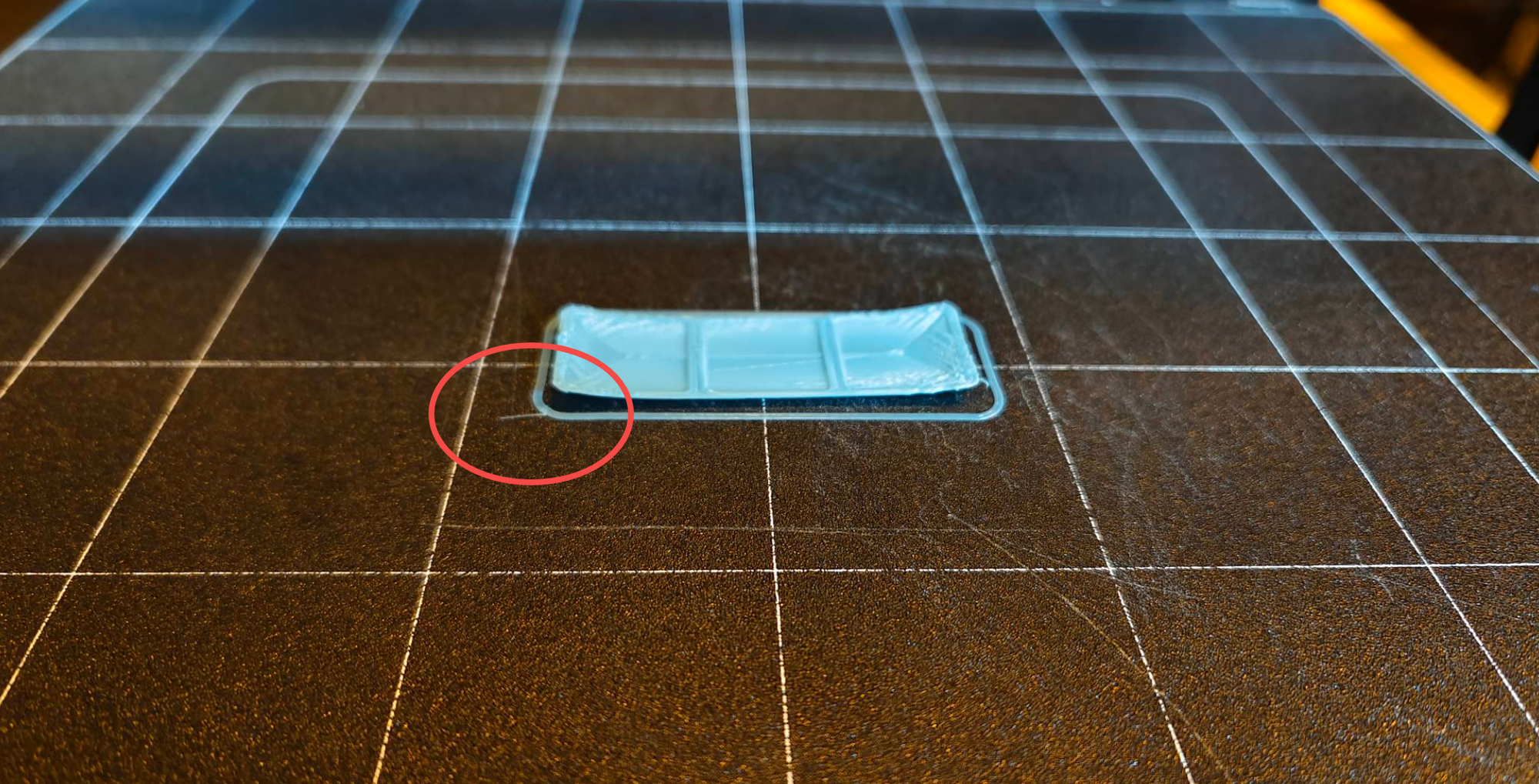 A close-up photograph of a blue 3D print on a textured print bed, with a red circle highlighting a corner that is warping and lifting off the surface despite the presence of a skirt.