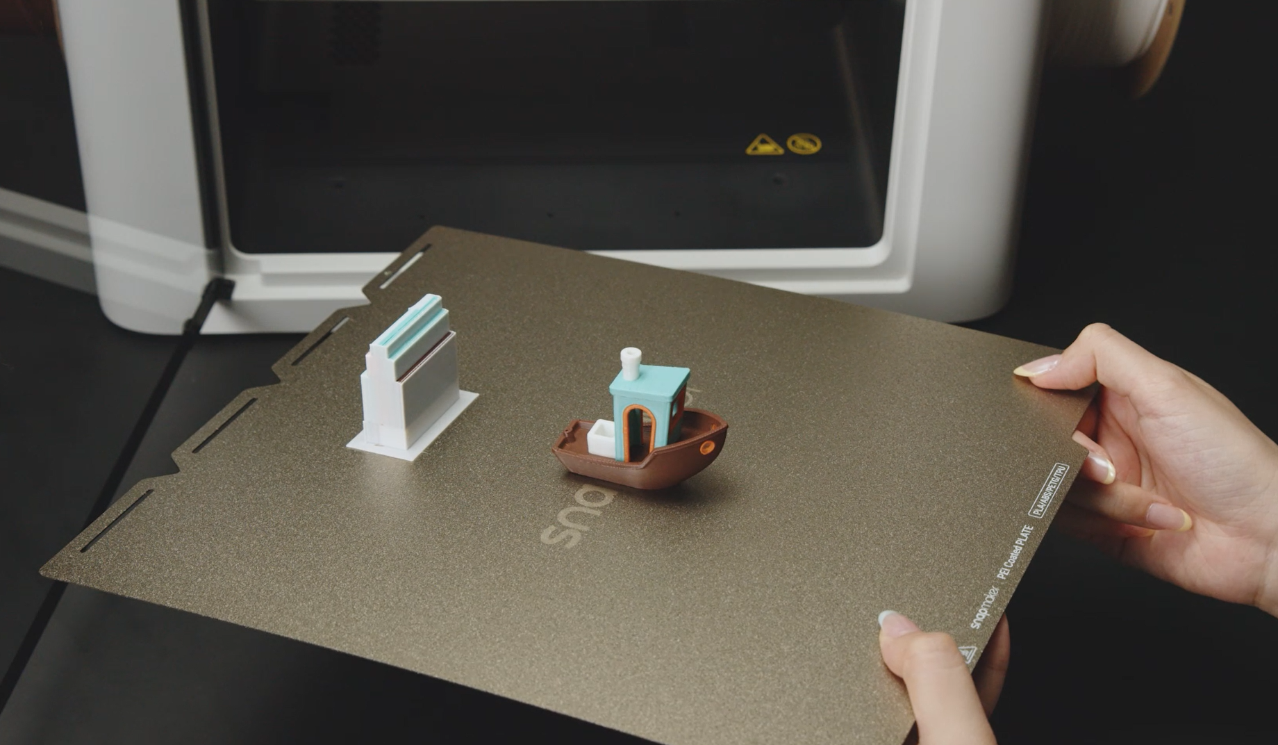 A person is holding a flexible PEI coated build plate from a Snapmaker 3D printer, featuring two multi-color printed models, including a Benchy boat.