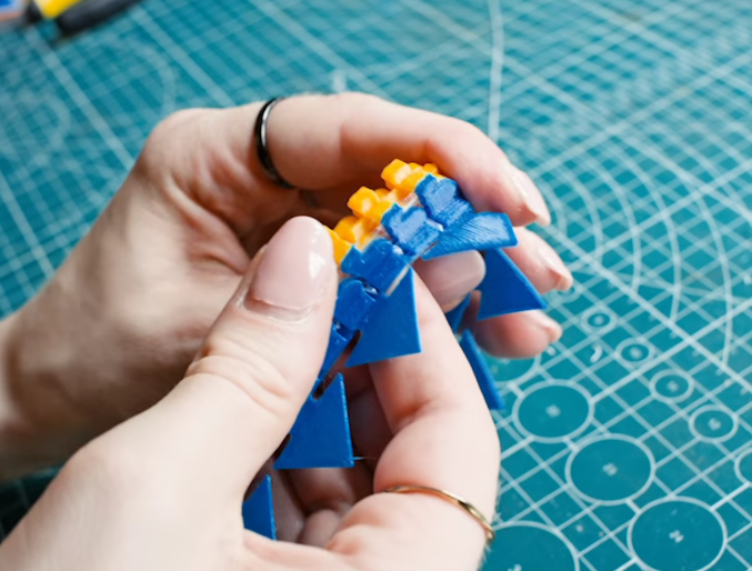 Hands bending a blue and yellow articulated 3D print, showcasing the capability to match flexible TPU and rigid PETG materials for creative, functional artwork.