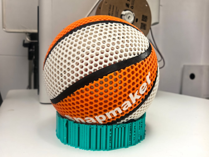 A hollow 3D printed basketball constructed from a white and orange hexagonal lattice, demonstrating how geometric shells drastically reduce filament usage while avoiding top-layer pillowing defects.