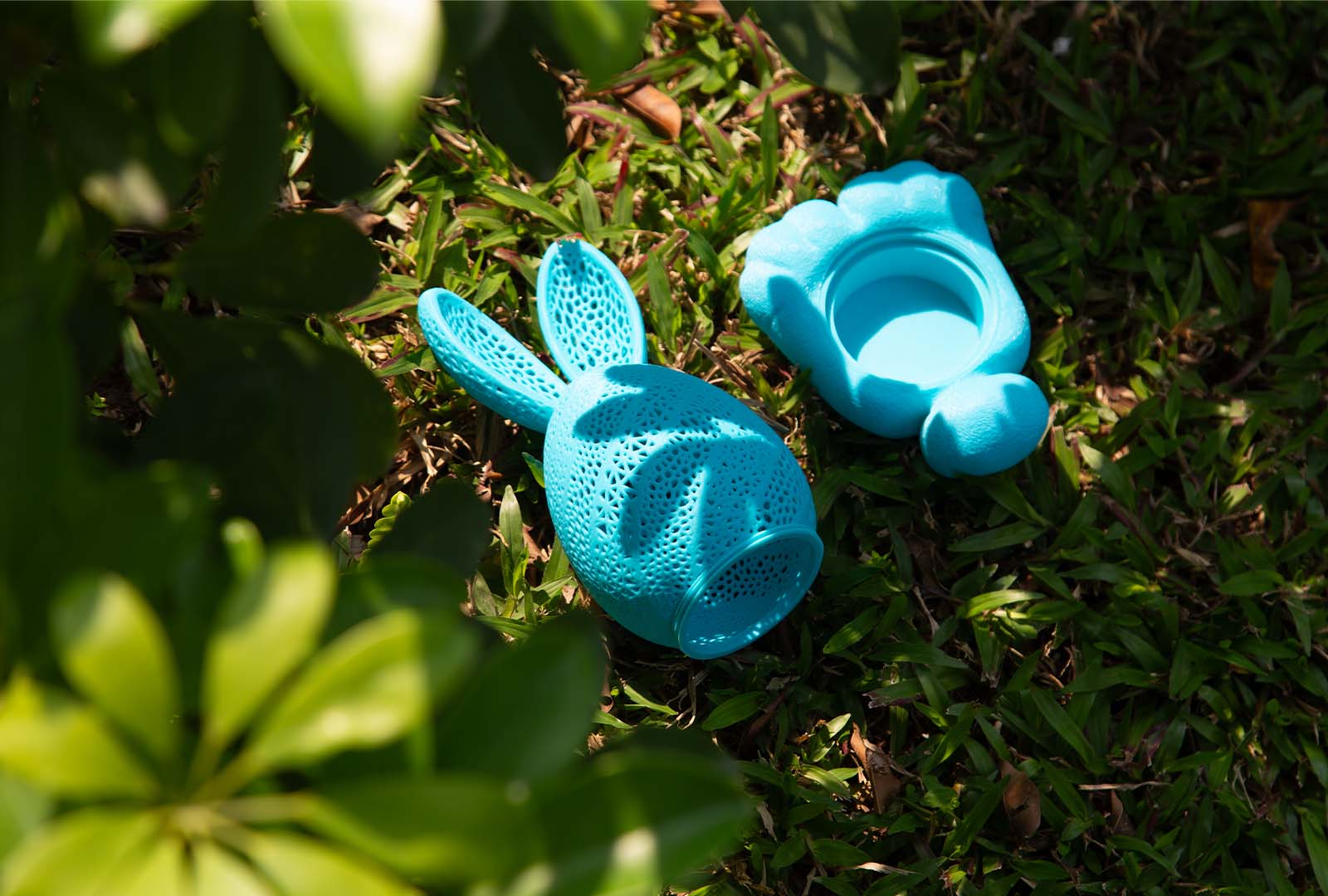 A bright cyan, intricately 3D printed hollow egg toy with a detailed porous lattice pattern and bunny-like features, resting on sunlit grass.