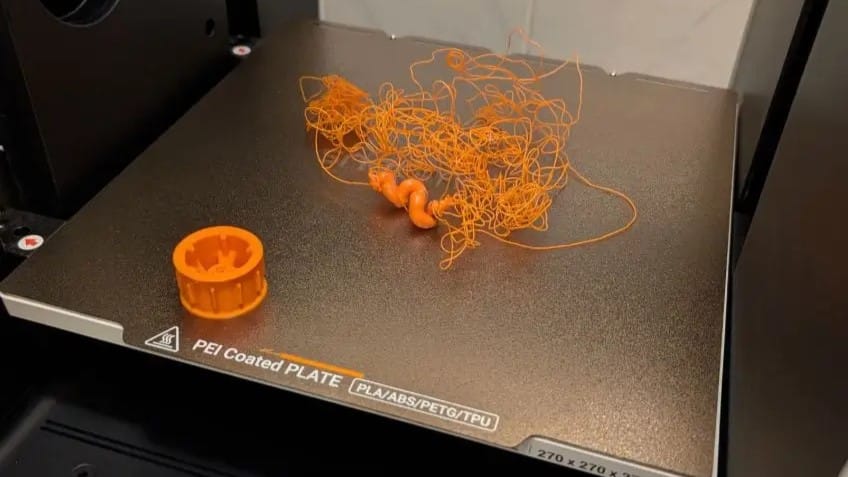 Failed 3D print showing tangled orange filament (spaghetti monster) on the PEI build plate, caused by filament feeding issues like tangled spools or grinding gears.