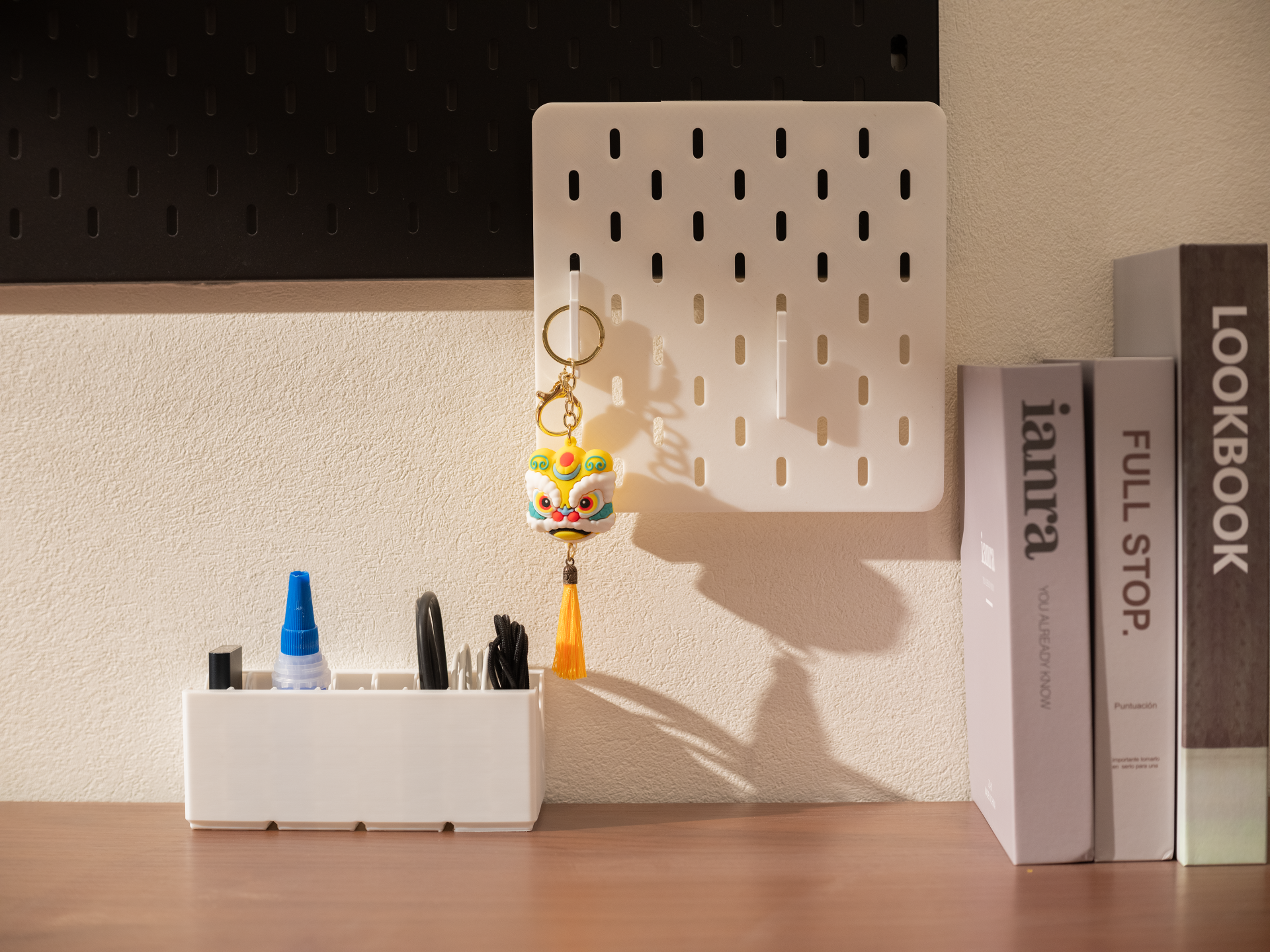 A clean workspace featuring a white 3D-printed slotted pegboard with a hanging keychain, positioned above a horizontal desk organizer holding tools and cables.