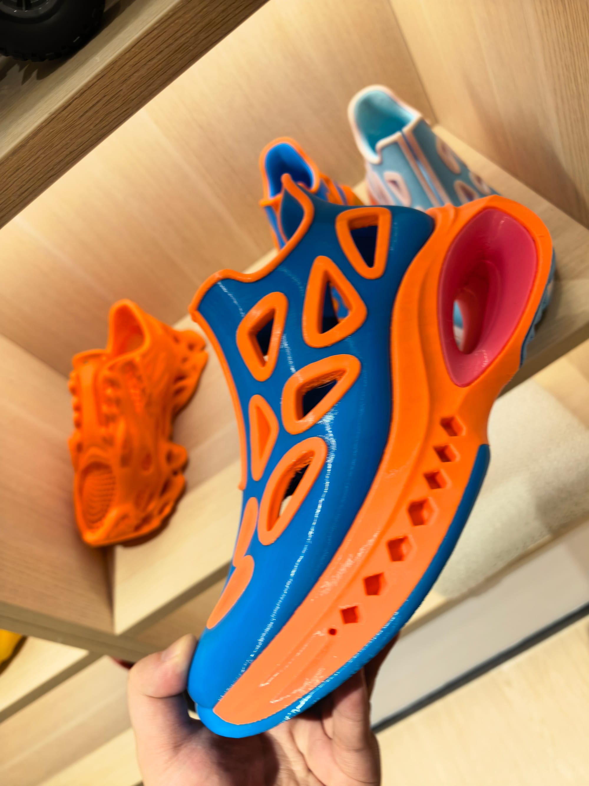 A close-up view of a person holding a blue and orange 3D-printed shoe to showcase the flexible filament material and visible print layer lines along the toe box.