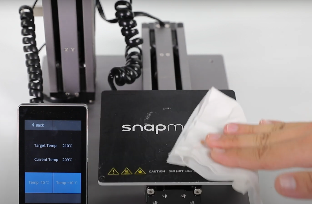 Wiping the PEI build plate of Snapmaker 3D printer, with nozzle temperature displayed on screen to solve poor first-layer adhesion and warping issues.