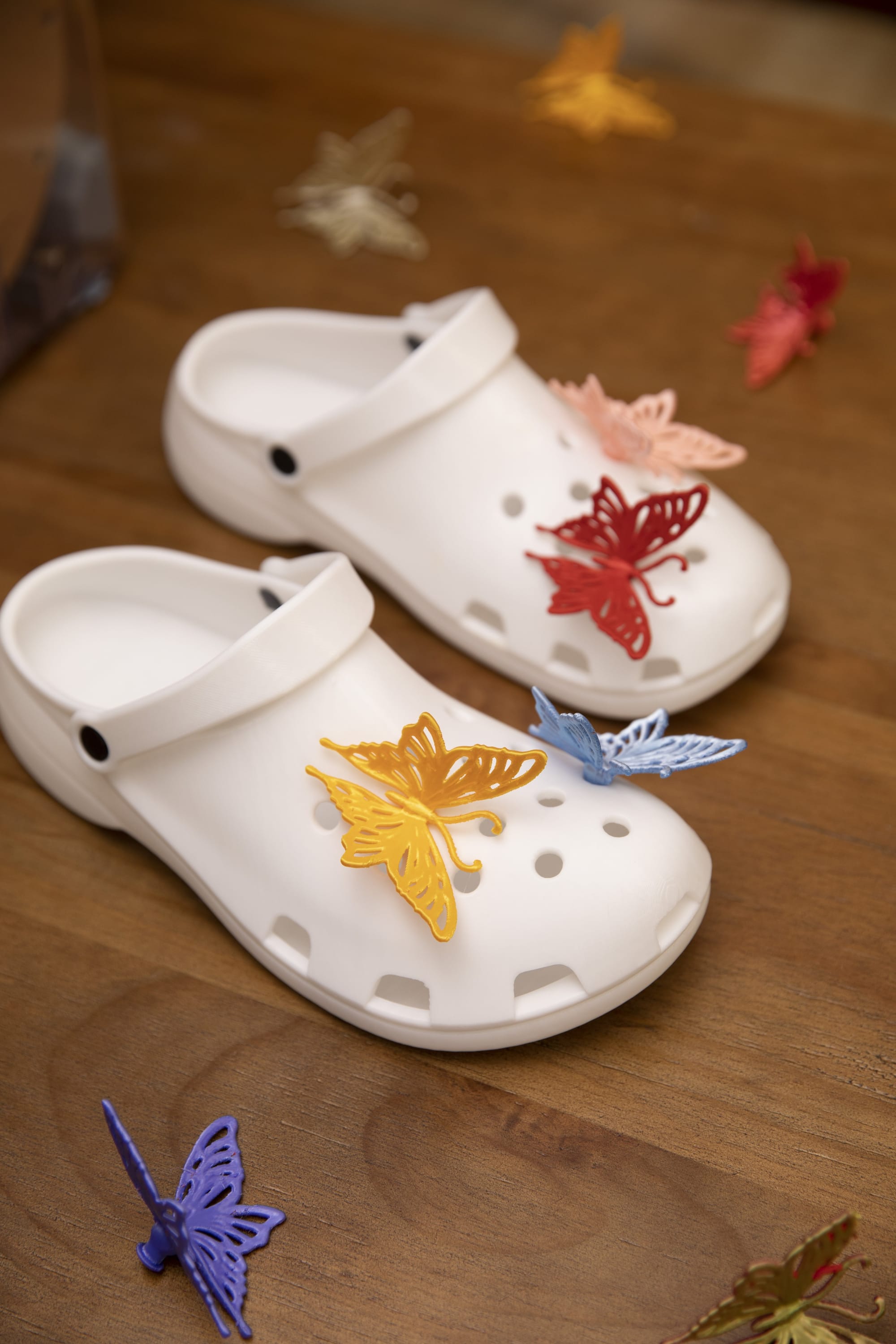 A pair of white 3D-printed clogs featuring various colorful butterfly charms arranged on a wooden surface.