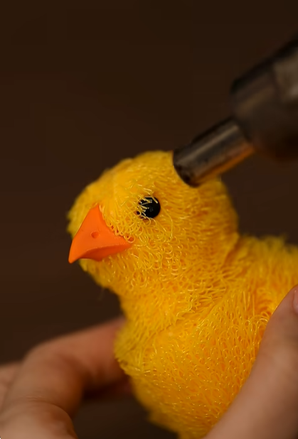 A heat gun being applied to a yellow 3D printed TPU chick to soften and style the chaotic extruded strands into a matted, fluffy finish.