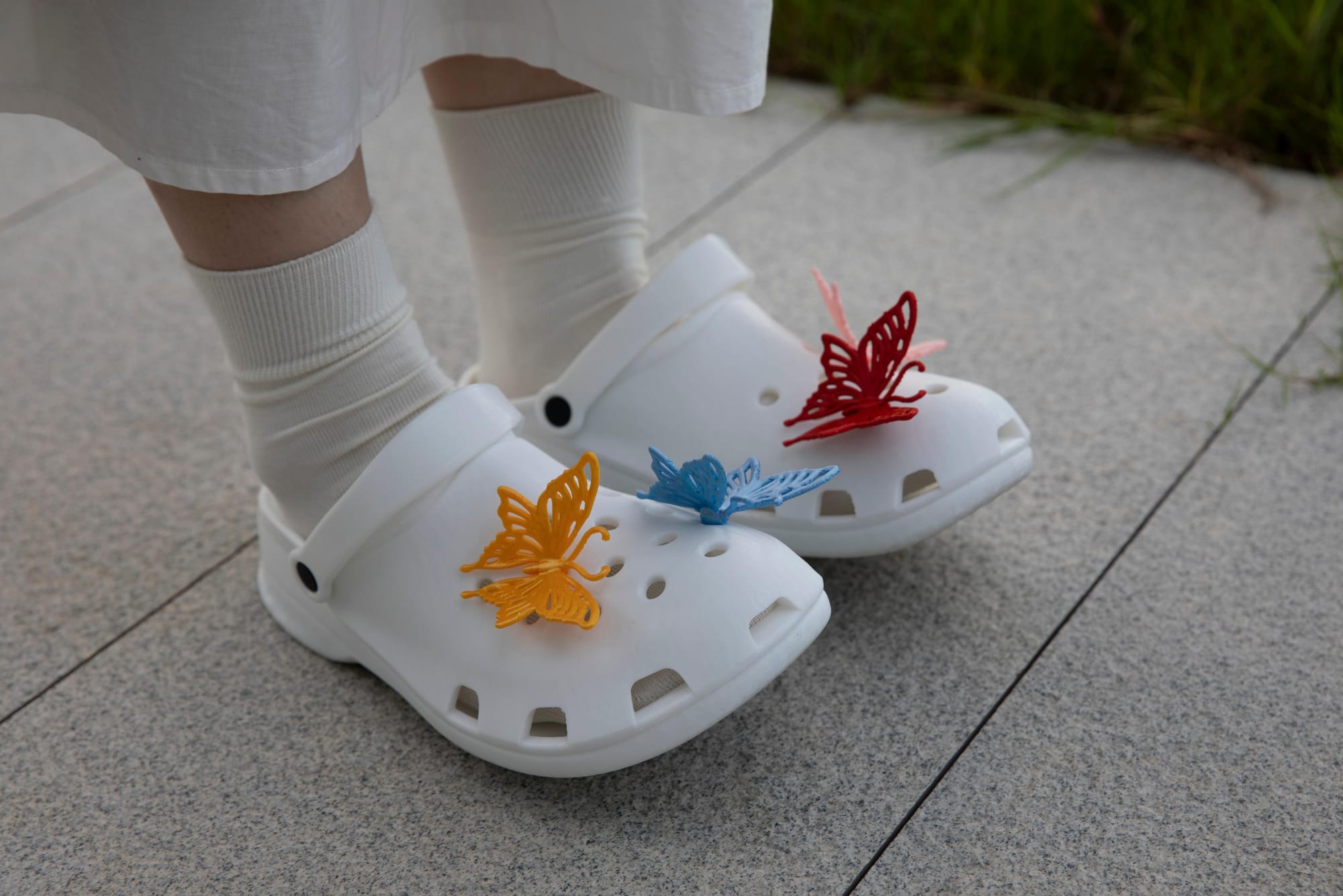 A person walking outdoors while wearing white 3D-printed TPU clogs accessorized with decorative butterfly charms.