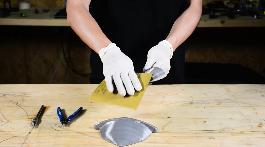 A person wearing protective gloves sands a grey 3D-printed part on a workbench to smooth the surface and prepare it for a protective coating.