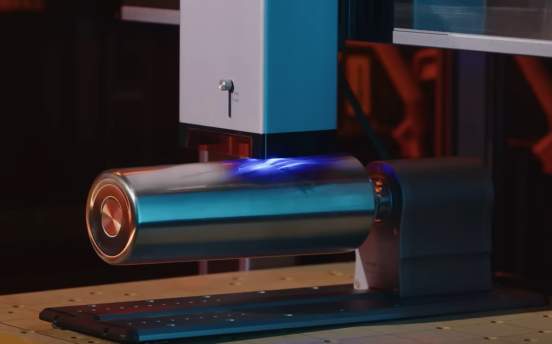 Laser machine engraving a design onto a silver cylindrical object, using a rotary attachment.
