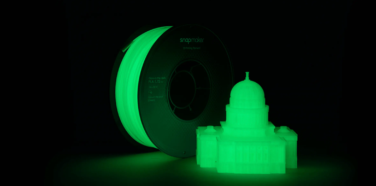 A spool of Snapmaker glow-in-the-dark filament and a 3D printed capitol building both glowing a vibrant green in a dark room.