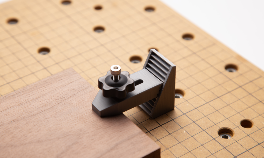 A close-up, angled shot of a gray 3D printed CNC jig with a knurled metal knob that is clamping a piece of wood to a grid-patterned bed.