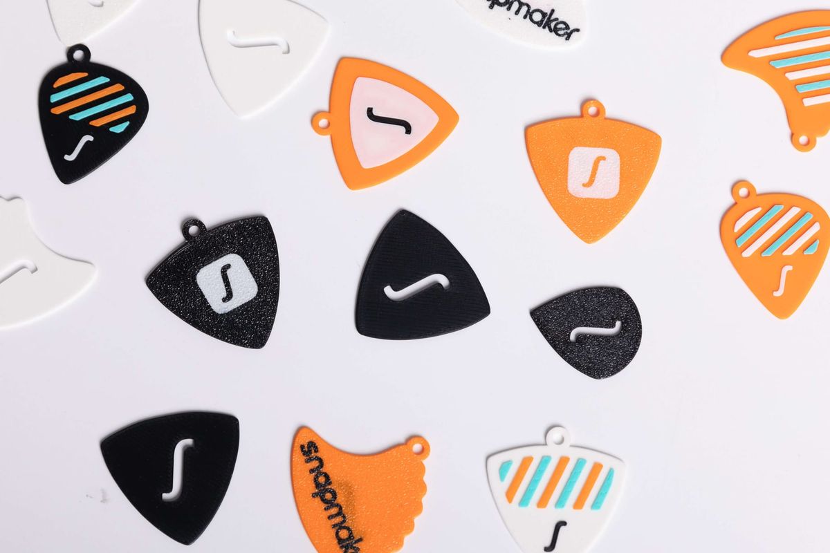 A scattered collection of various 3D-printed guitar picks in white, black, and orange, showcasing different shapes, patterns, and the Snapmaker logo.