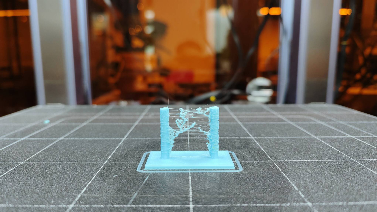 A close-up of a cyan 3D print retraction test on a build plate showing severe stringing and branching between the two towers.