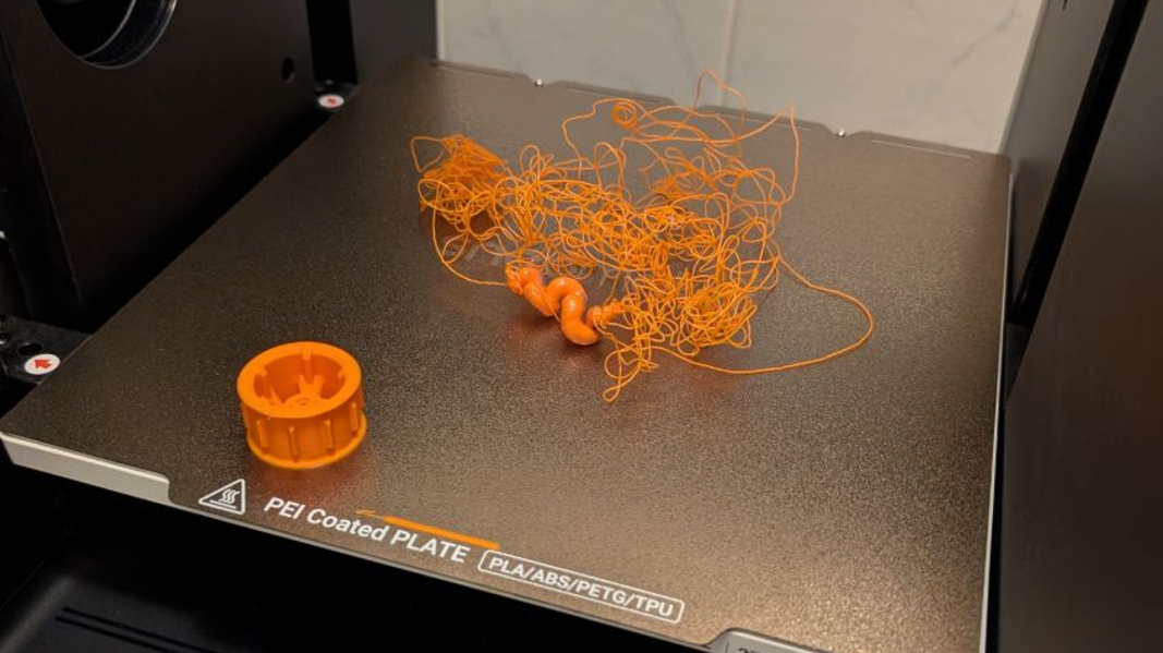 A failed 3D print on a PEI coated build plate showing a tangled "spaghetti" mess of orange filament, a common symptom of poor bed adhesion caused by incorrect nozzle height.