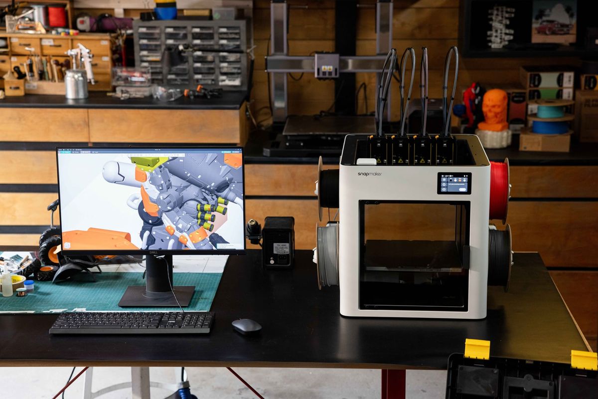 A Snapmaker U1 3D printer sits on a workbench next to a computer monitor displaying pre-processing slicing software, workflow from digital mesh translation to physical hardware execution.