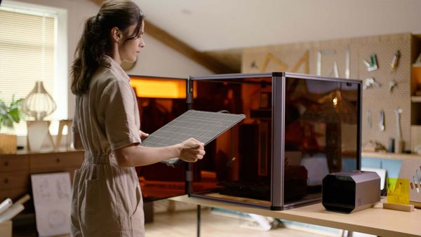 A woman in a beige jumpsuit holds a build plate while performing maintenance on a large enclosed 3D printer in a well-lit, modern workshop.