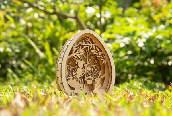 A laser-cut layered Easter egg decor made of wood, showcasing bunnies, trees, and leaves in a three-dimensional design, placed on a grassy field with a blurred green background.