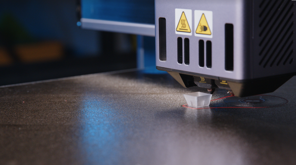 A macro shot of a 3D printer nozzle extruding a translucent PETG filament to print a small keycap on a textured build plate, illustrating the FDM printing process.
