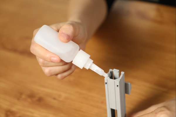 A person carefully applies glue from a bottle into a joint of a gray part during the assembly process.