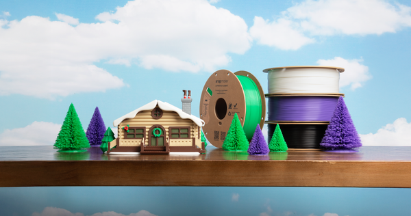 A small, detailed 3D printed model of a log cabin with miniature trees, arranged as a diorama on a wooden surface with a backdrop of blue sky and clouds.